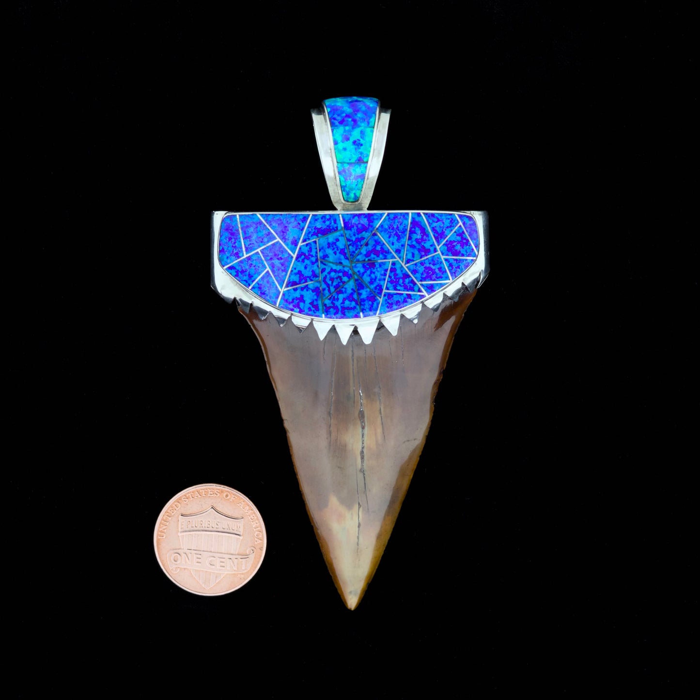 Fossil mako shark tooth pendant with blue inlay and silver setting on a black background, next to a penny for scale.